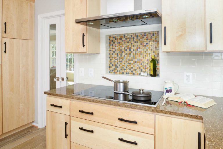light wood cabinetry modern kitchen with electric stovetop and stainless steel hood vent mosaic tile backsplash detail