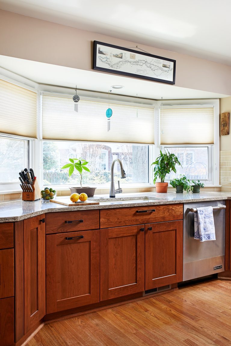 kitchen sink in front of large bay window medium stained cabinetry and wood floors