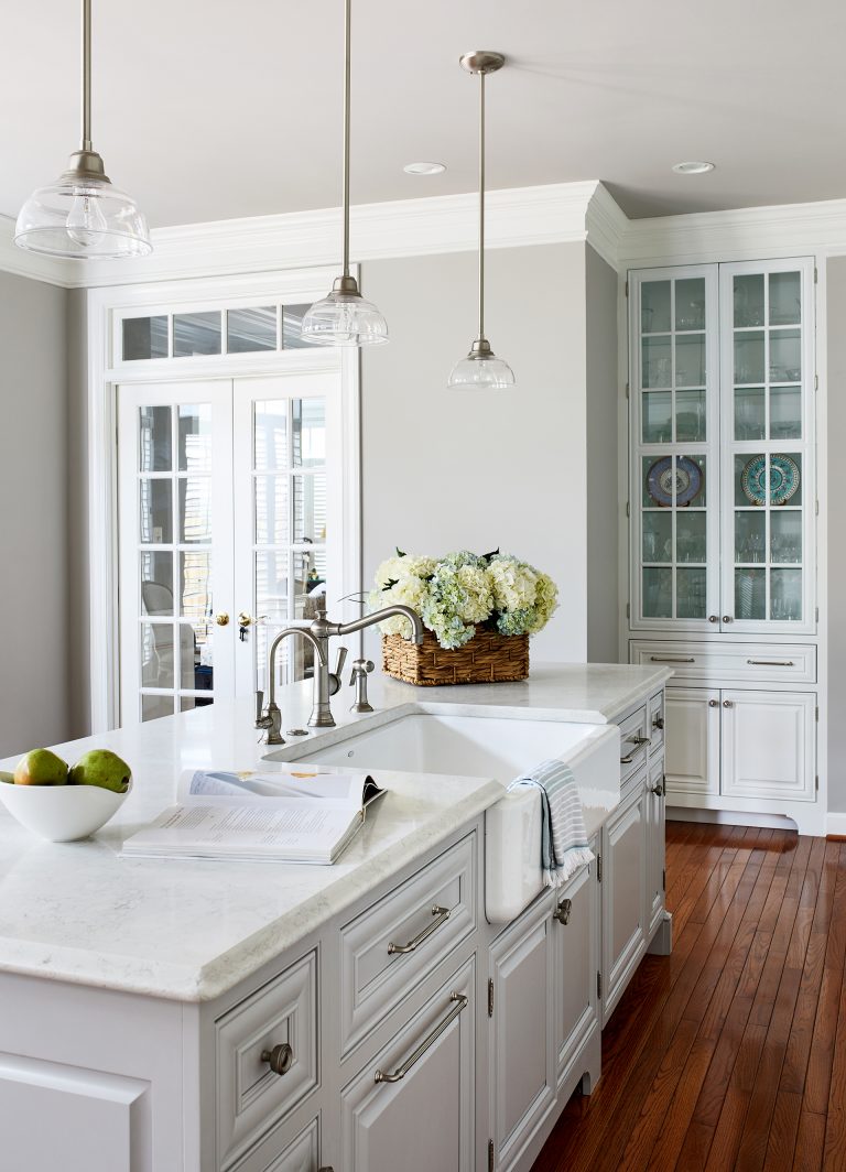 traditional kitchen with 3- hanging light pendants over kitchen island, large white farmhouse sink, hardwood floors