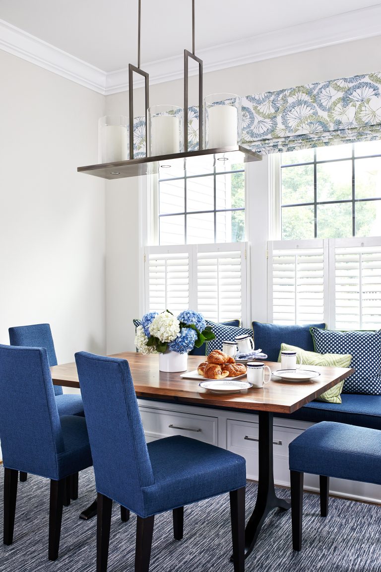blue kitchen breakfast nook features a built-in bench window seat topped with blue cushion and blue pillows under a bay window facing a wood dining table with blue cushion dining chairs