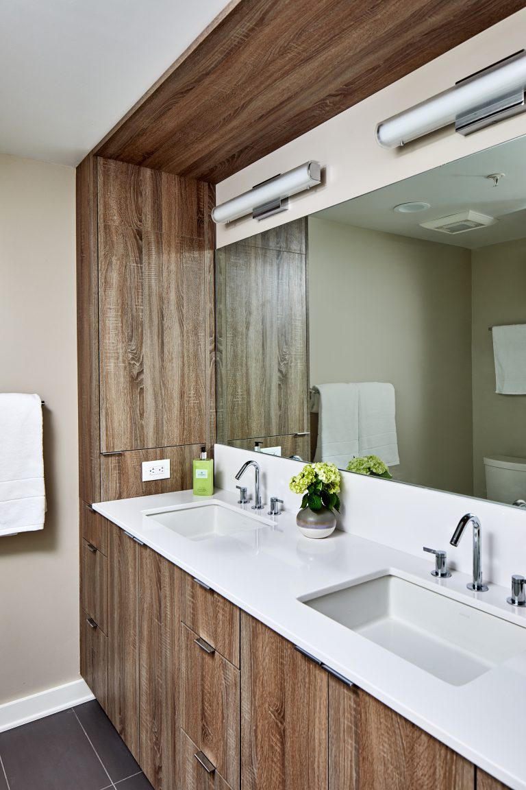 Remodeling company bathroom with wide mirror that runs length of vanity with wood cabinets with pull handles and white rectangular sinks