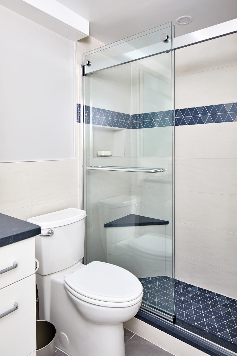 walk-in shower with frameless shower glass door with blue triangle tile on shower floor and middle of the shower wall with a small sitting seat